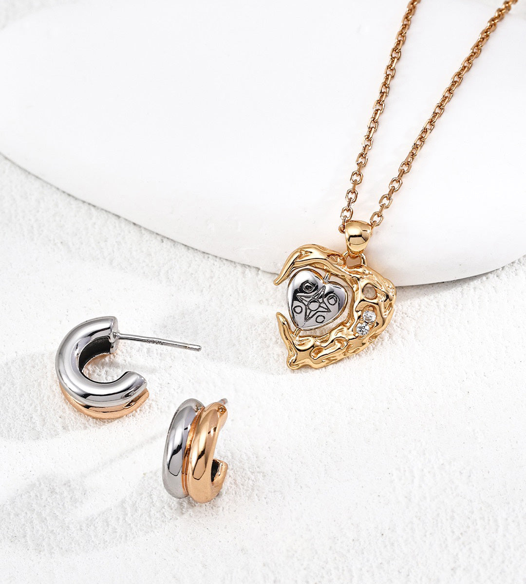 A jewelry set featuring a gold-plated pendant necklace with a heart-shaped design and matching two-tone hoop earrings in gold-plated and white-gold plated, displayed on a white textured surface.