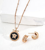 An 18K gold-plated on S925 silver jewelry set featuring a round pendant necklace with a black center and an 18K gold-plated Medusa head, accompanied by matching gold-plated hoop earrings, displayed on a white textured surface with a white block background.