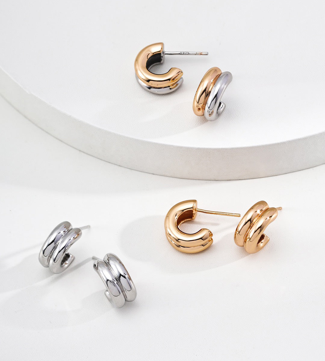 Three pairs of stacked hoop stud earrings, arranged on a clean white surface with a curved white pedestal.