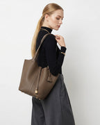 A woman in a black ribbed turtleneck and dark gray wide-leg trousers carries an Extra Large Togo Leather Bucket Tote Bag in Elephant Grey Color with a gold-toned lock