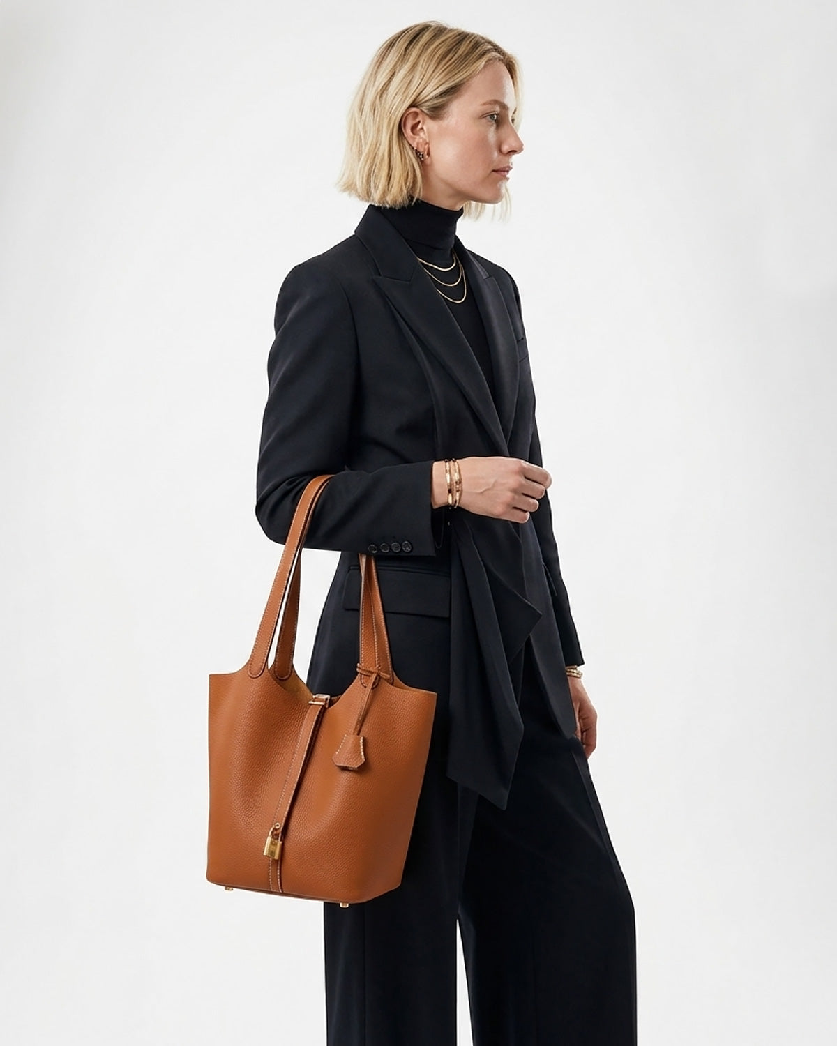 A woman in a black turtleneck, blazer, and trousers carries an Extra Large Togo Leather Bucket Tote Bag in Light Brown Color with Gold-toned Hardware and Lock
