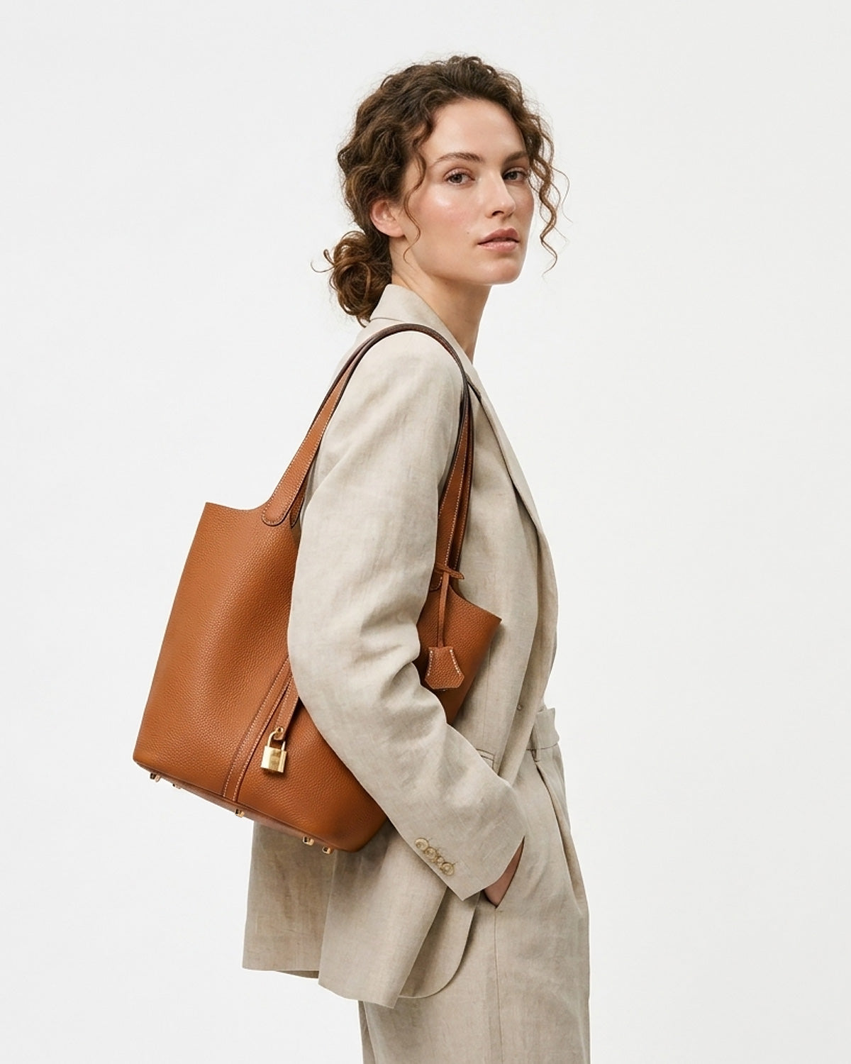 A woman in a beige blazer carries a Large Togo Leather Bucket Tote Bag in Light Brown Color with Gold-toned Hardware and Lock