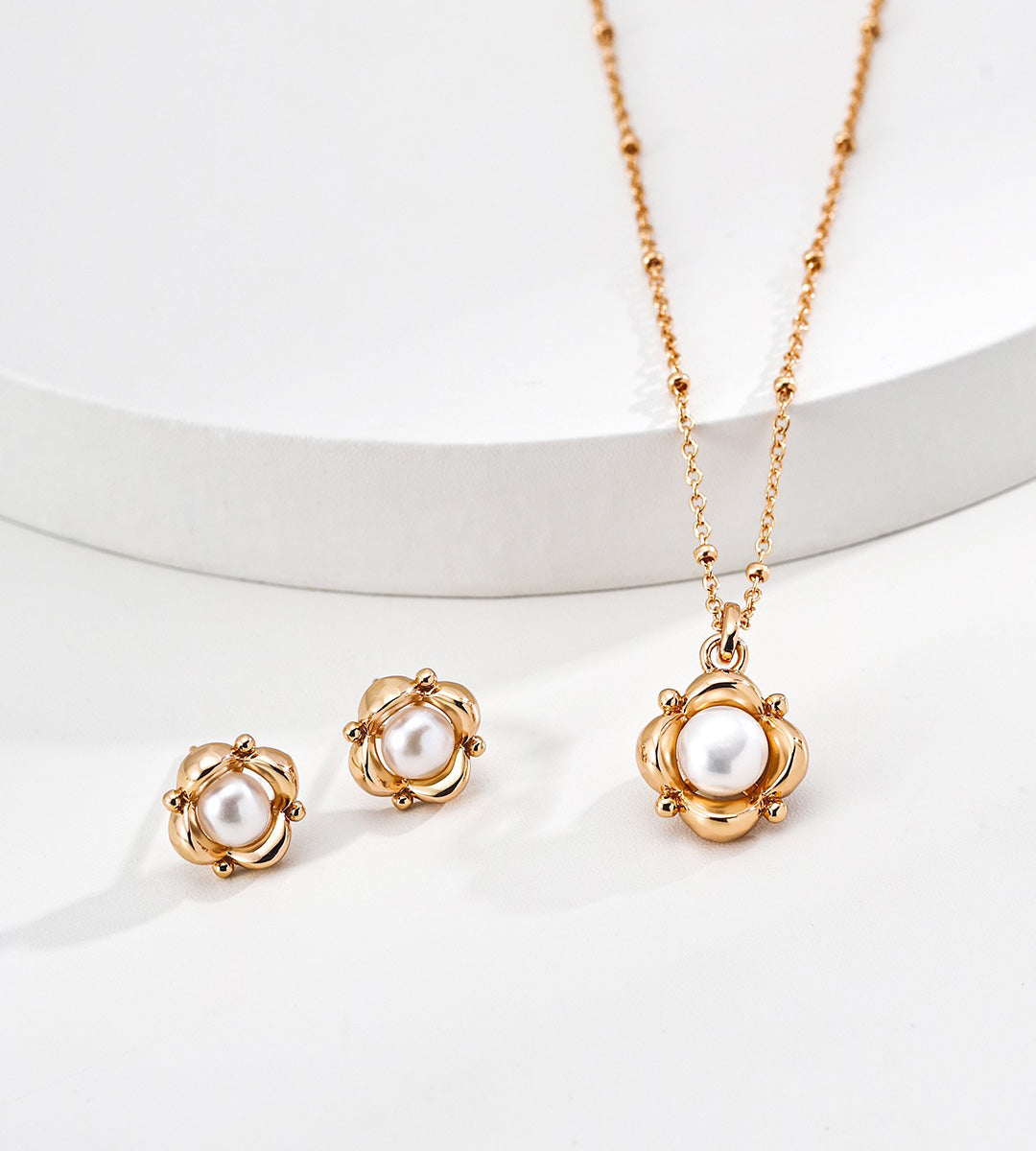 18K gold-plated necklace and matching stud earrings set, each featuring a round white pearl surrounded by a textured floral-like gold frame, displayed on a clean white surface with a curved white pedestal.
