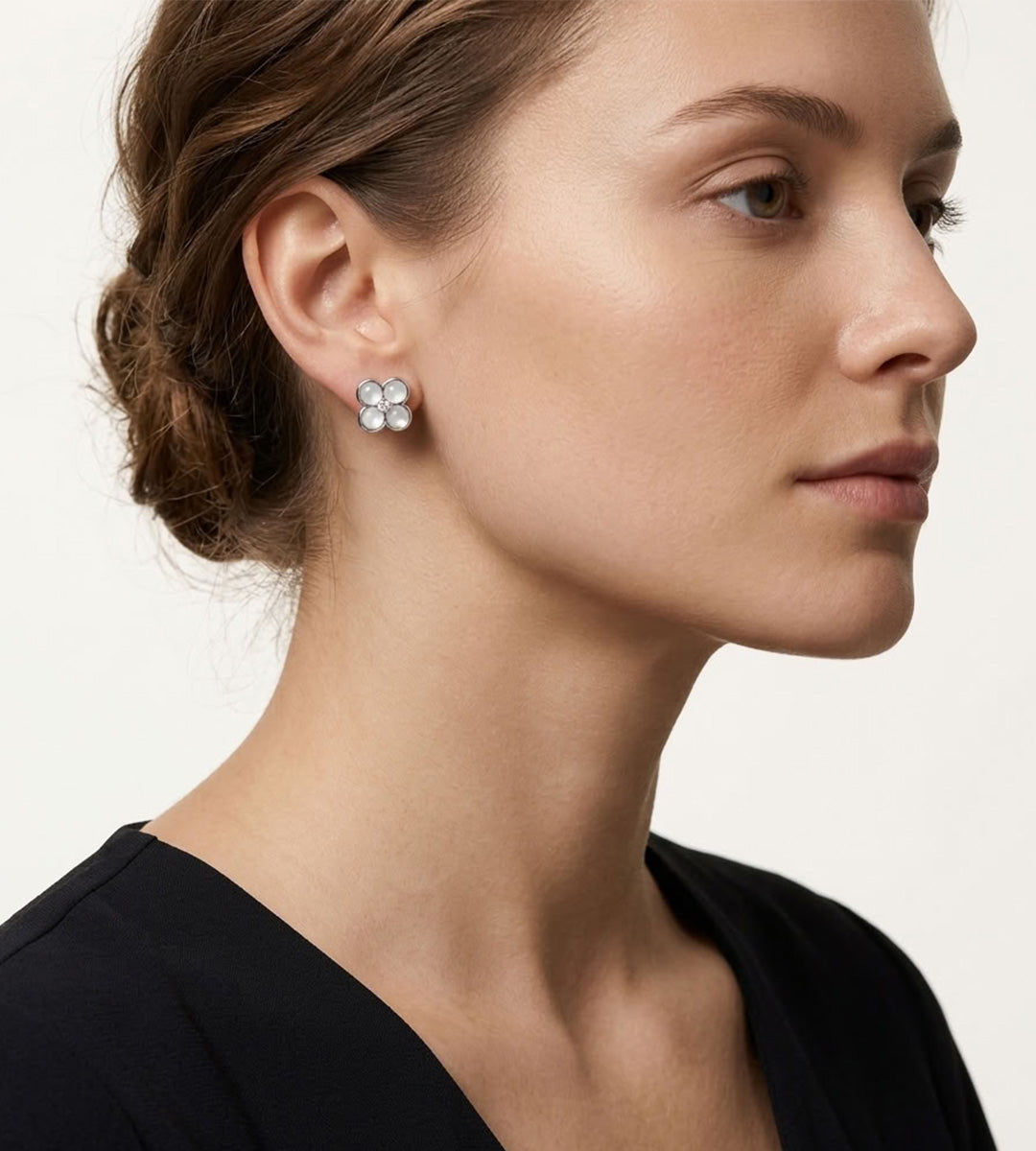 A close-up side profile of a woman wearing a small silver-toned flower-shaped stud earring with clear stones, against a plain light background.