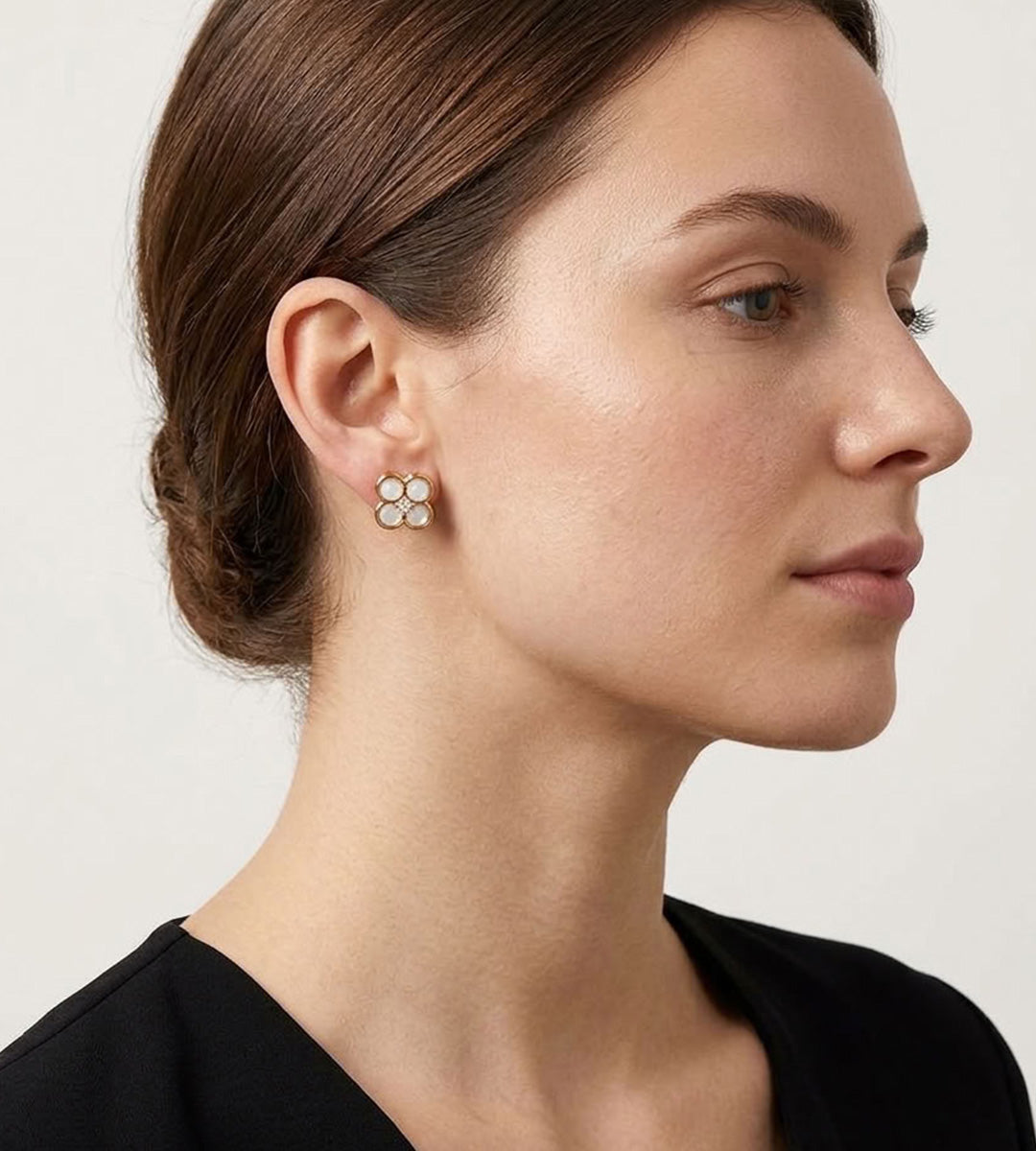 A close-up side profile of a woman with brown hair in a low bun, wearing a small gold stud earring with four white pearl-like beads, against a clean off-white background.