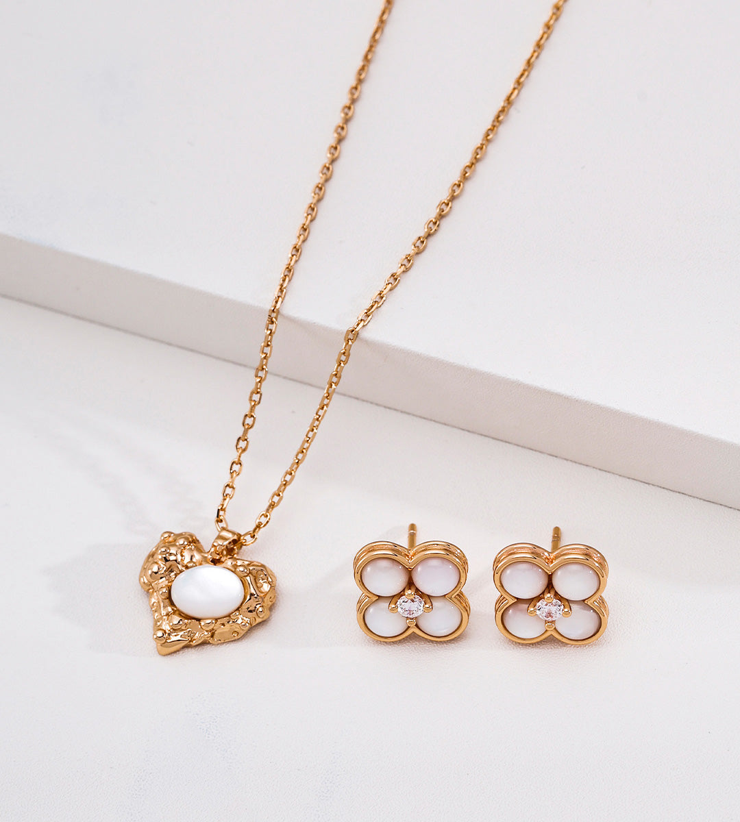 An 18K gold-plated S925 silver necklace and matching stud earrings displayed on a clean white surface. The necklace has a textured gold heart-shaped pendant with a white oval center, and the 18K gold-plated S925 silver earrings are four-petal clover shapes with white centers and small cubic zirconia stones.