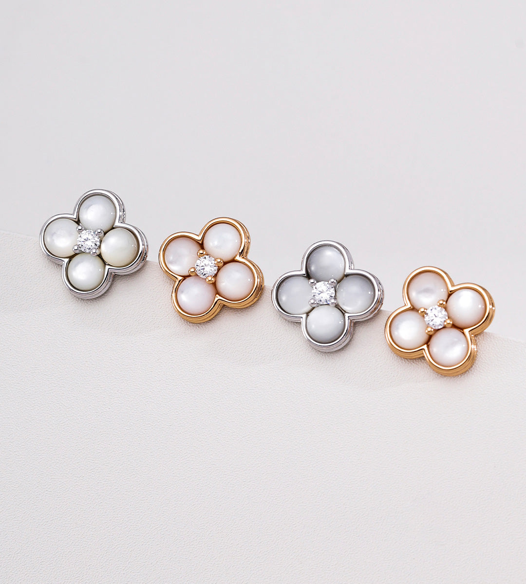 Four flower-shaped stud earrings arranged in a row on a light gray background. Each earring features five white pearl-like round stones set in a gold or silver-toned metal border, with a small clear center stone.