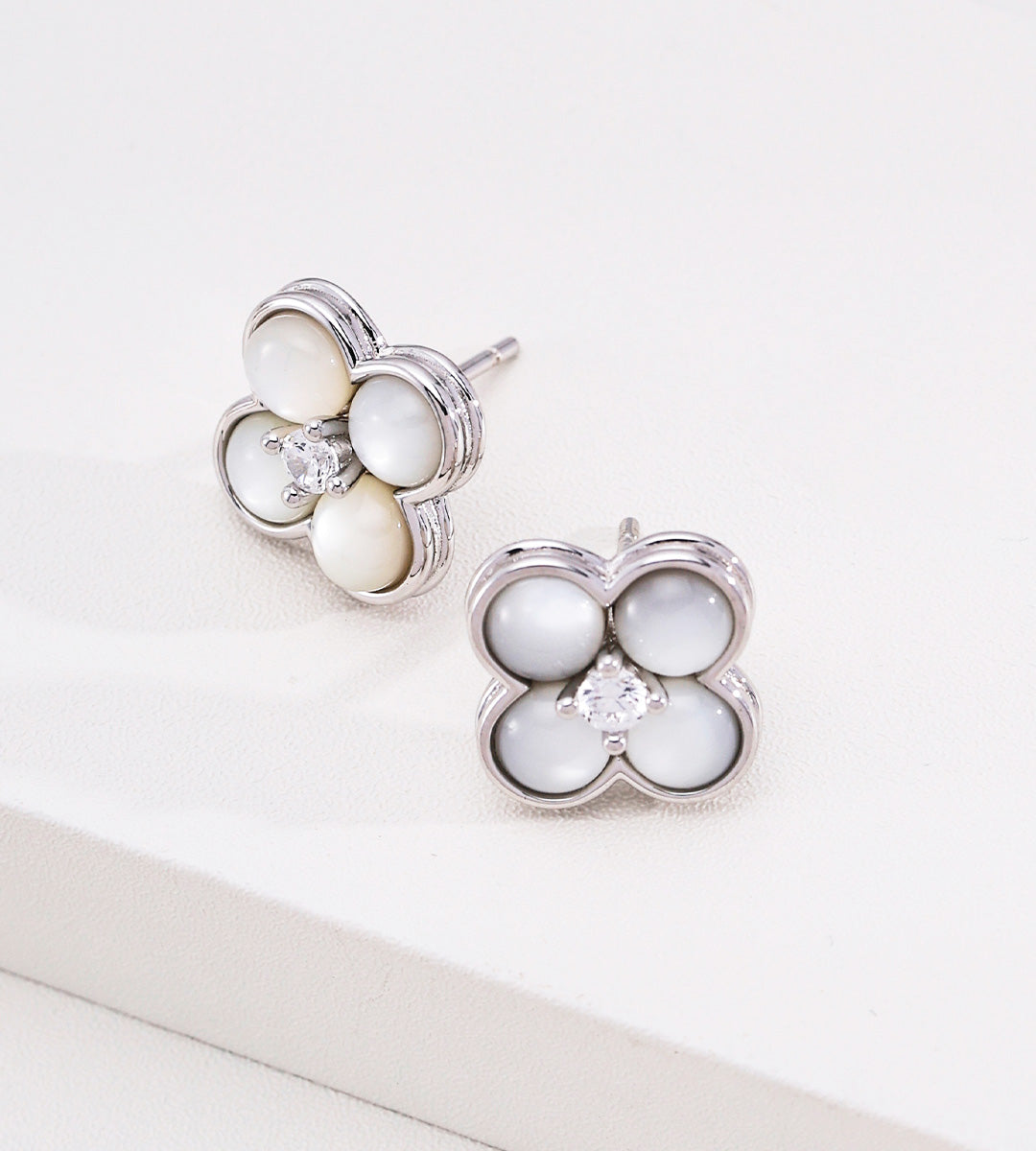 A pair of 18K white-gold-plated S925 silver four-petal flower stud earrings with white mother-of-pearl inlays and a small cubic zirconia at the center, displayed on a clean white surface.