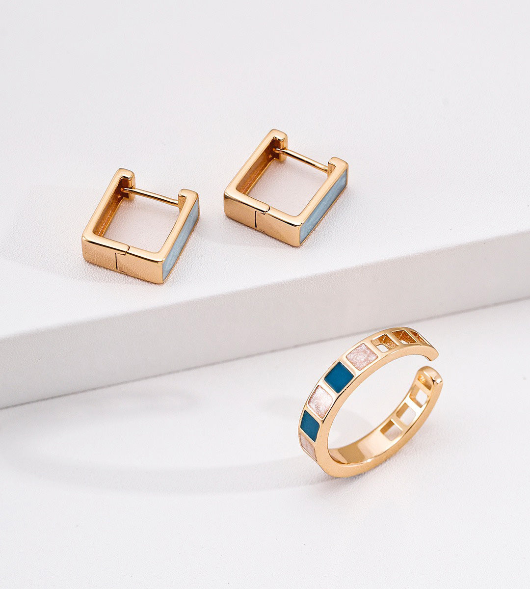 A pair of 18K gold-plated square hoop earrings and a matching gold open ring with alternating light blue and pale pink square inlays, displayed on a clean white surface.