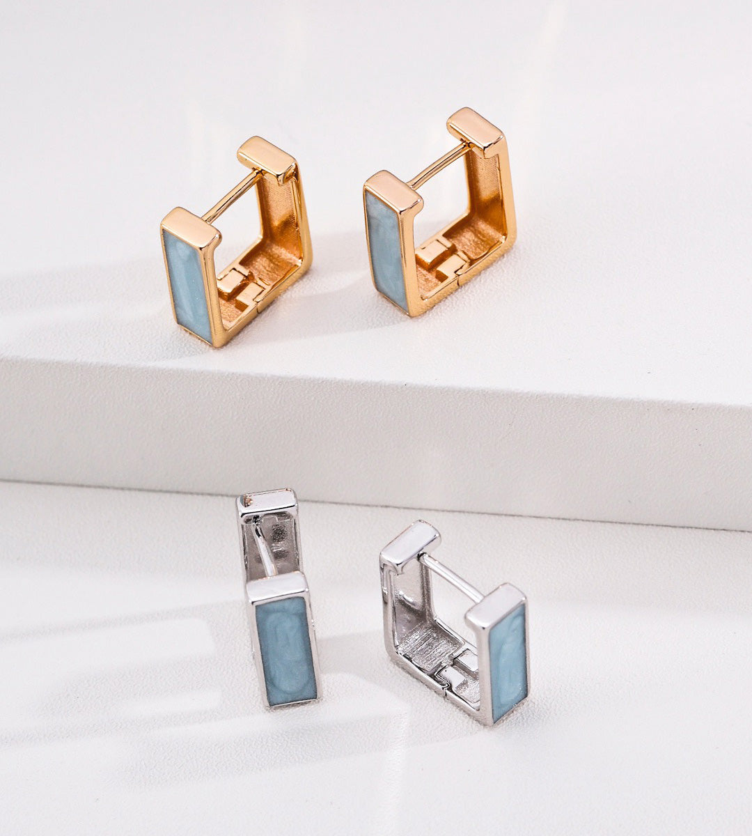 Four geometric square stud earrings displayed on a clean white surface, showing both gold and silver metal finishes with light blue inlay panels.