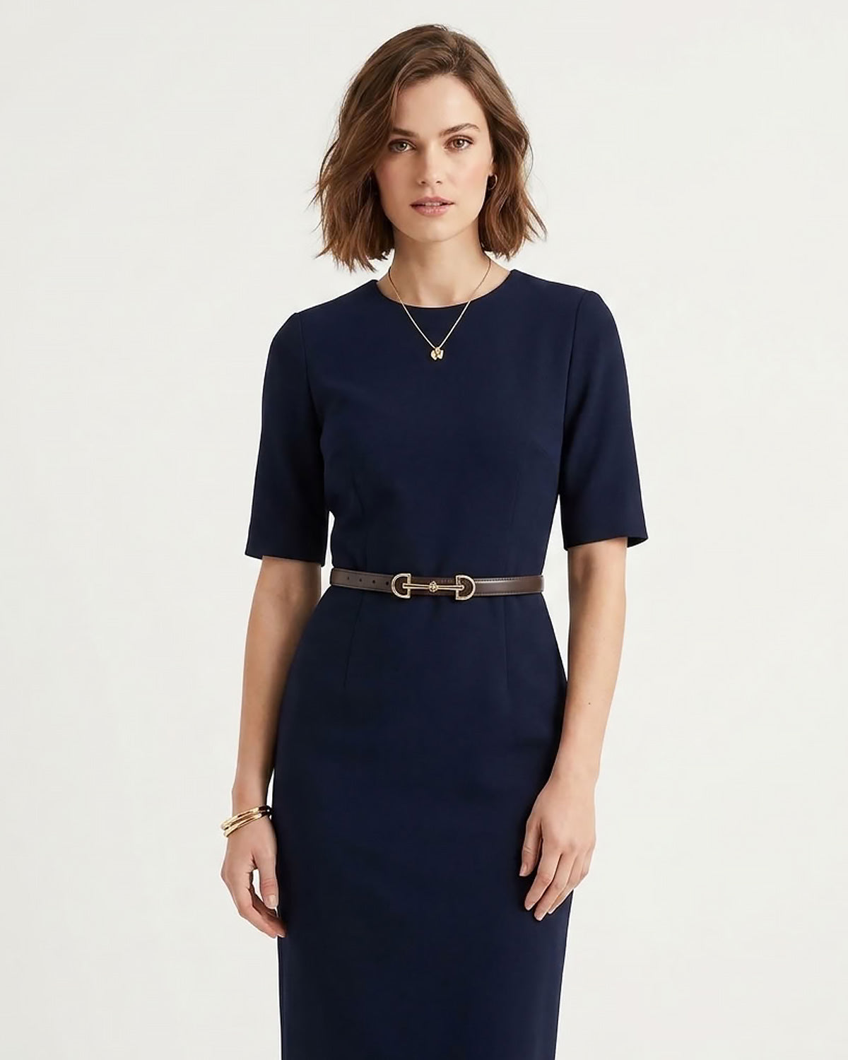 A woman modeling a navy blue short-sleeve dress with a thin classic brown belt featuring a gold buckle, set against a plain light background.