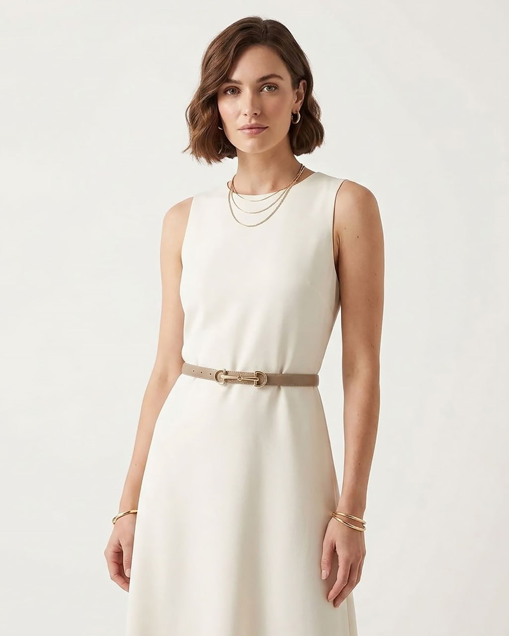 A woman wearing a sleeveless cream-colored dress with a thin tan classic belt with a gold horsebite buckle at the waist, styled with layered gold necklaces and bracelets.