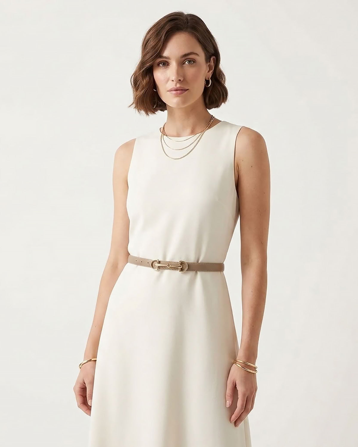 A woman wearing a sleeveless cream-colored dress with a thin tan classic belt with a gold horsebite buckle at the waist, styled with layered gold necklaces and bracelets.