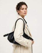 A woman in a cream trench coat carries a black leather slouchy shoulder bag with knotted strap details | BUCKLE Collective