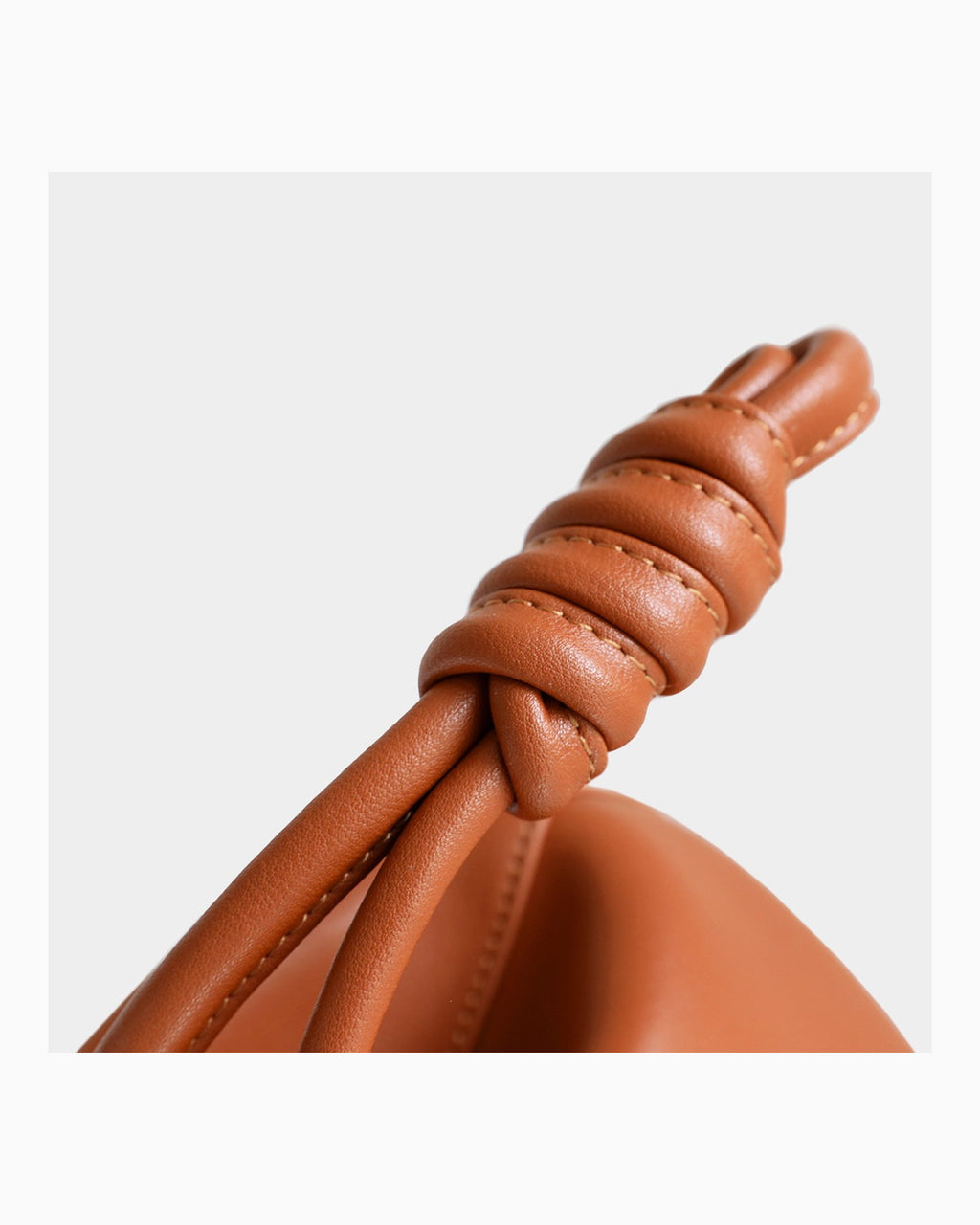 Close-up of a leather strap with a knotted detail and visible stitching, attached to a rounded tan leather product against a light gray background.