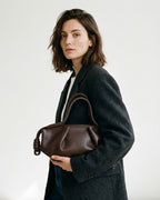 A woman wearing a dark charcoal wool coat and white shirt holds a dark brown leather shoulder bag by BUCKLE Collective