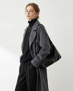 A woman models a dark gray wool-blend coat over a black turtleneck and black trousers, carrying a large black slouchy Hobo bag against a plain light gray studio background.