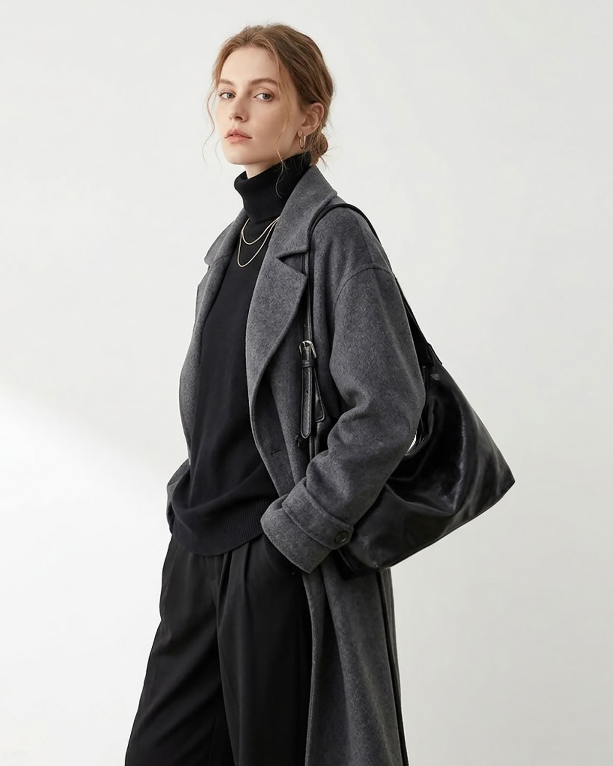 A woman models a dark gray wool-blend coat over a black turtleneck and black trousers, carrying a large black slouchy Hobo bag against a plain light gray studio background.
