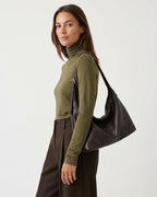 A woman wearing an olive green turtleneck and dark brown trousers, carrying a large dark brown genuine leather slouchy hobo shoulder bag against a plain light gray studio background.