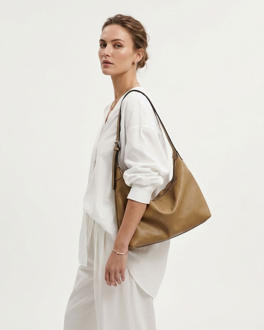 A woman in a white outfit models a large taupe leather slouchy Hobo bag against a plain white studio background.