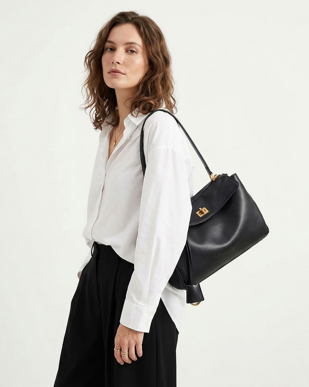A woman wearing a white button-down shirt and black skirt carries a black leather shoulder bag with flap closure and gold twist-lock clasp against a plain off-white background.