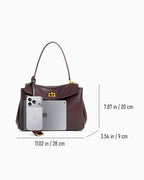 A dark burgundy leather handbag shown on a light gray background with overlaid dimensions and a phone with tablet for scale