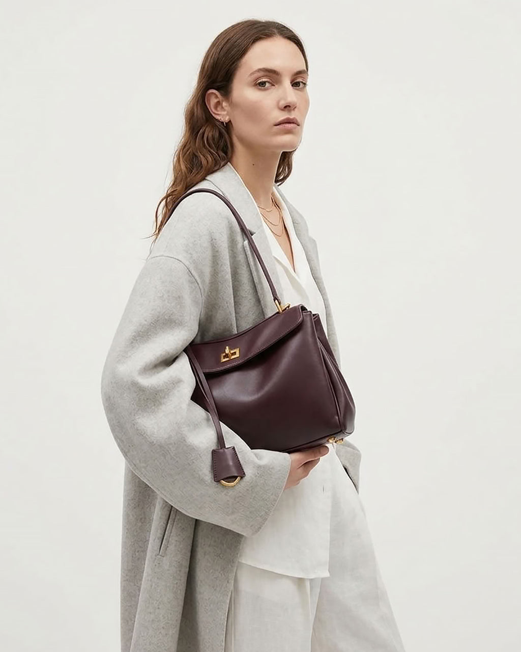 A woman wearing a light gray coat and white top carries a dark burgundy leather shoulder bag with gold hardware against a plain off-white background.