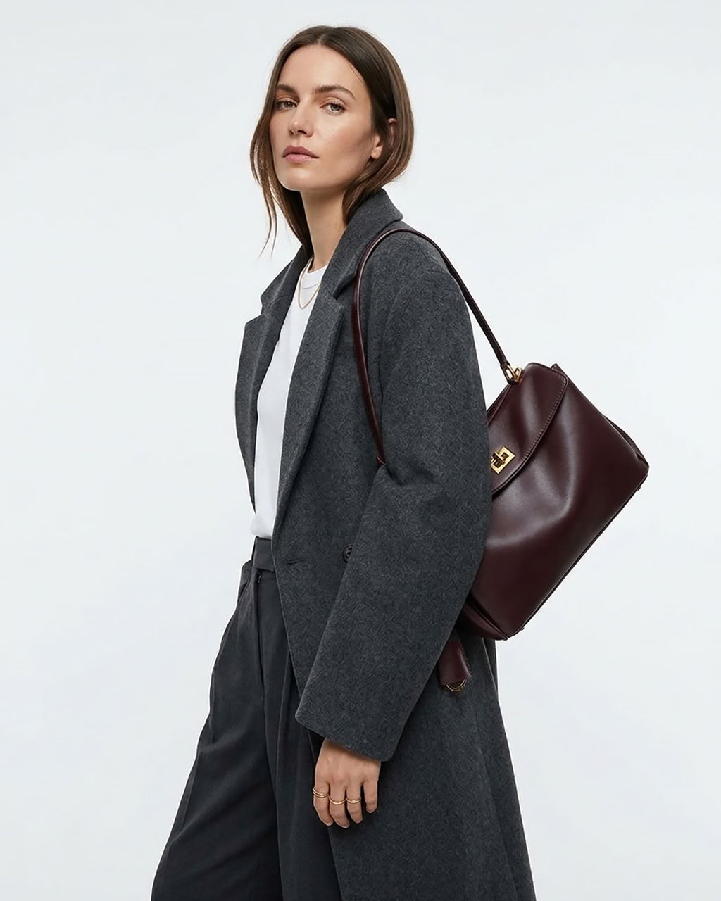 A woman wearing a dark gray wool coat over a white top, carrying a burgundy leather shoulder bag with gold hardware against a light gray studio background.