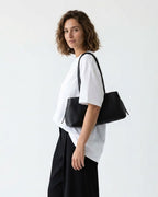 A woman with short brown hair is shown in profile wearing a white oversized T-shirt, black wide-leg pants, and carrying a black leather shoulder bag | BUCKLE Collective