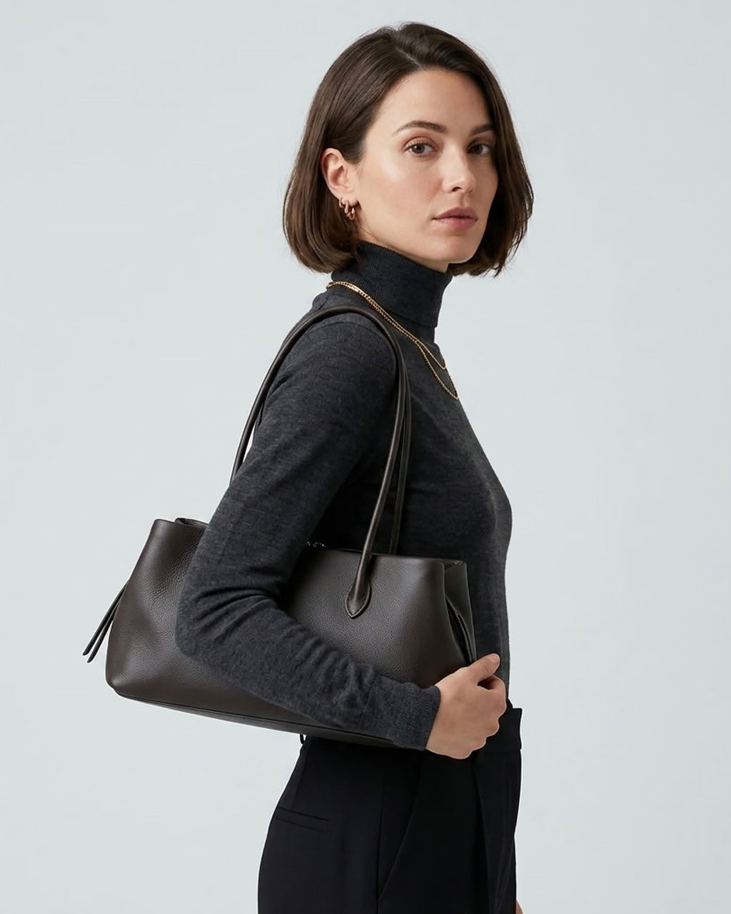 A woman with a short brown bob hairstyle is shown from the side, wearing a dark charcoal turtleneck sweater and black trousers while carrying a Dark Coffee Brown Genuine Full-Grain Leather Commuter Shoulder Bag by BUCKLE Collective