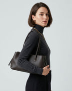 A woman with a short brown bob hairstyle is shown from the side, wearing a dark charcoal turtleneck sweater and black trousers while carrying a Dark Coffee Brown Genuine Full-Grain Leather Commuter Shoulder Bag by BUCKLE Collective