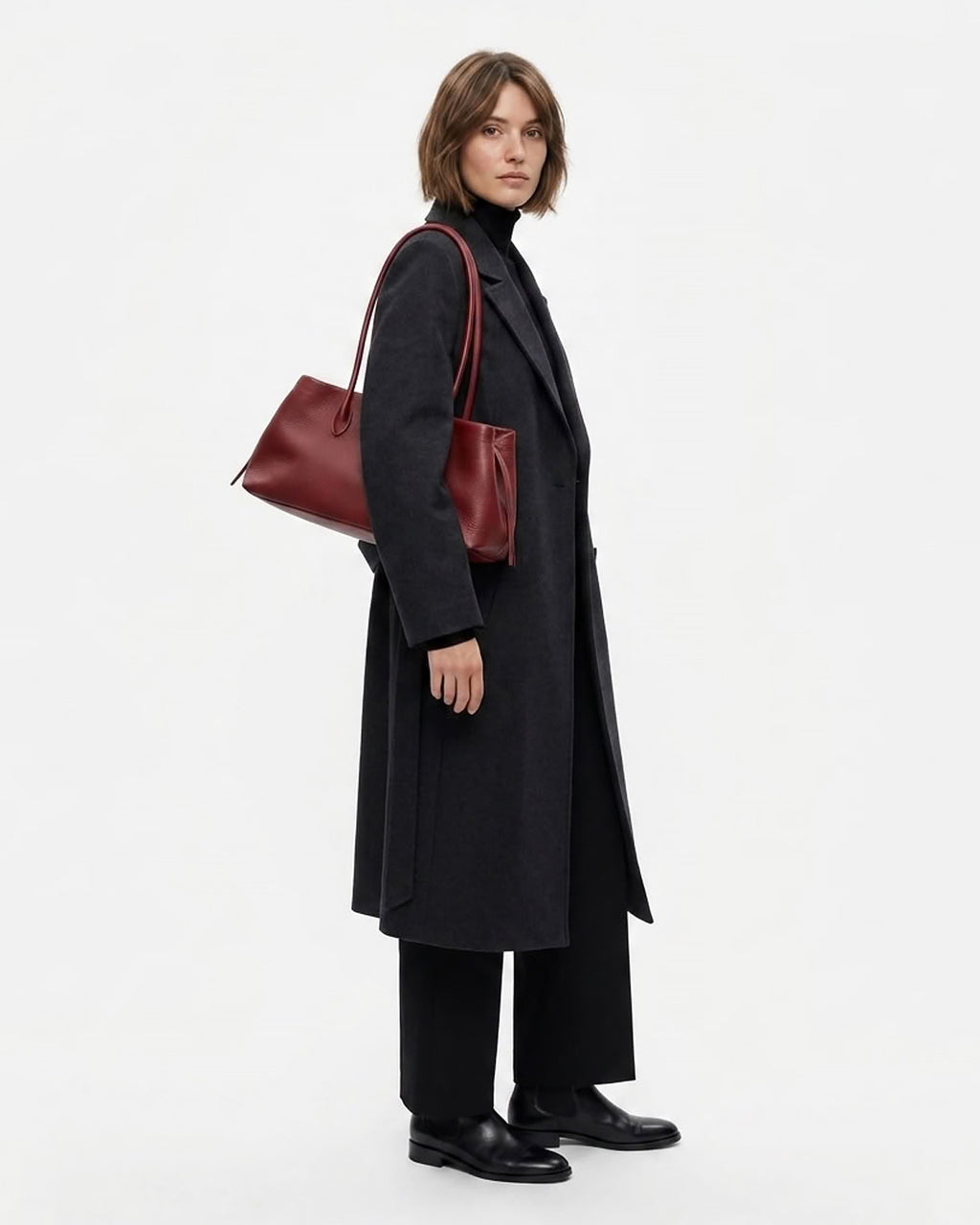 A model wearing a long black coat, black trousers, and black shoes, carrying a dark red leather shoulder bag by BUCKLE Collective