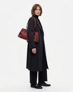 A model wearing a long black coat, black trousers, and black shoes, carrying a dark red leather shoulder bag by BUCKLE Collective