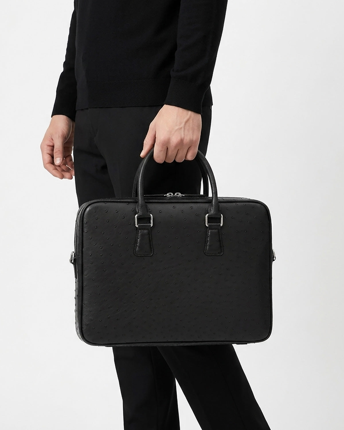 A man in a black outfit holding a black genuine ostrich leather briefcase with top handles and silver-tone hardware. BUCKLE Collective