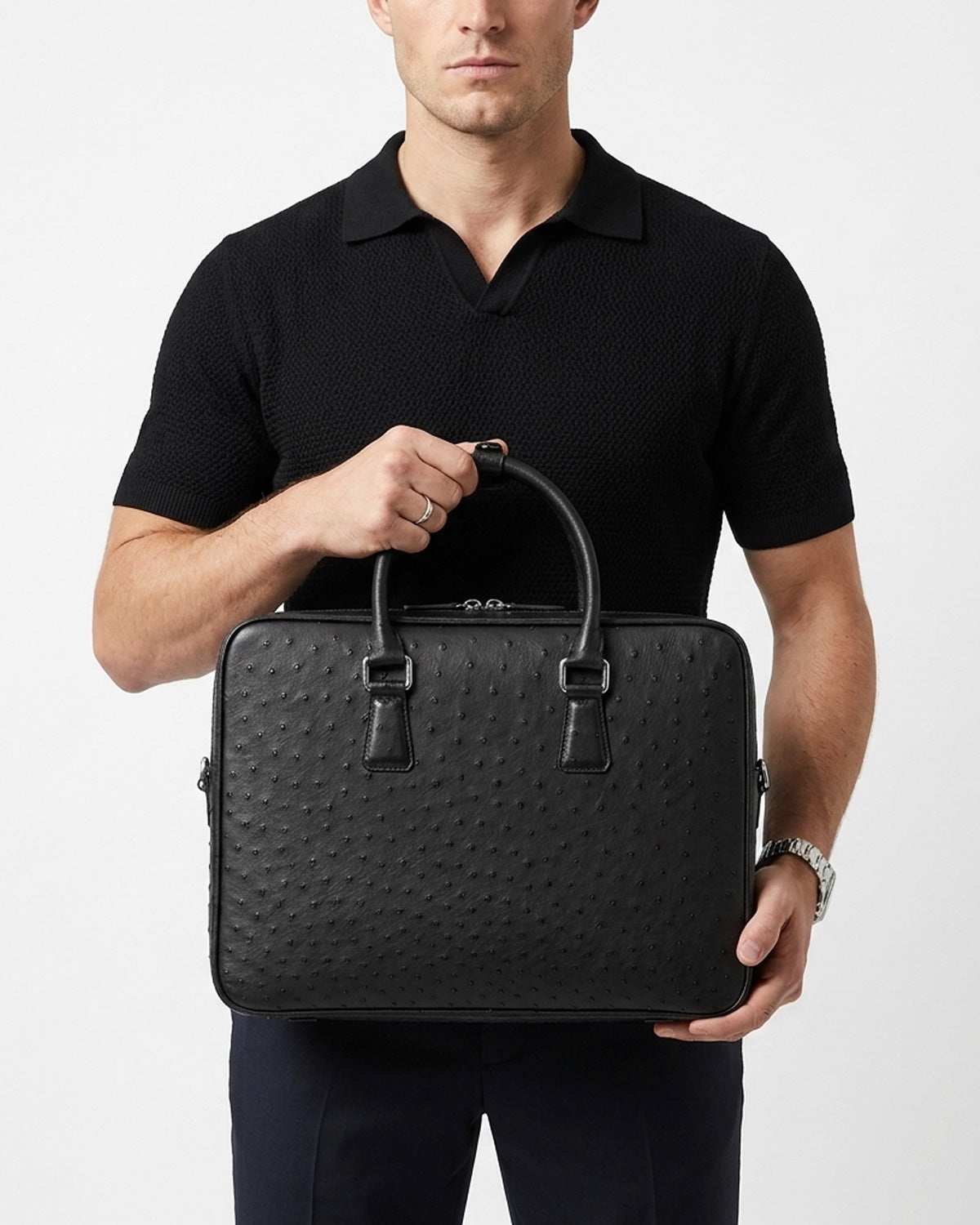 A man wearing a black polo shirt holds a black genuine ostrich leather briefcase by BUCKLE Collective