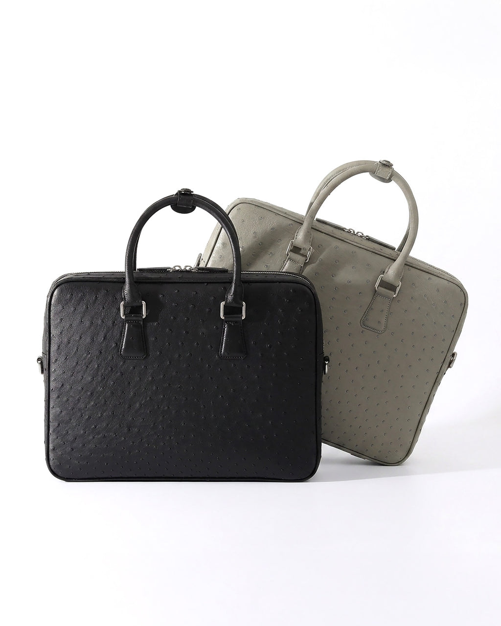 Two structured leather briefcases with top handles, shown on a clean white studio background. The front bag is a black genuine ostrich briefcase, and the rear bag is an elephant gray genuine ostrich briefcase | BUCKLE Collective