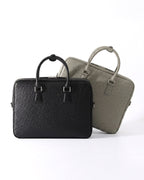 Two structured leather briefcases with top handles, shown on a clean white studio background. The front bag is a black genuine ostrich briefcase, and the rear bag is an elephant gray genuine ostrich briefcase | BUCKLE Collective