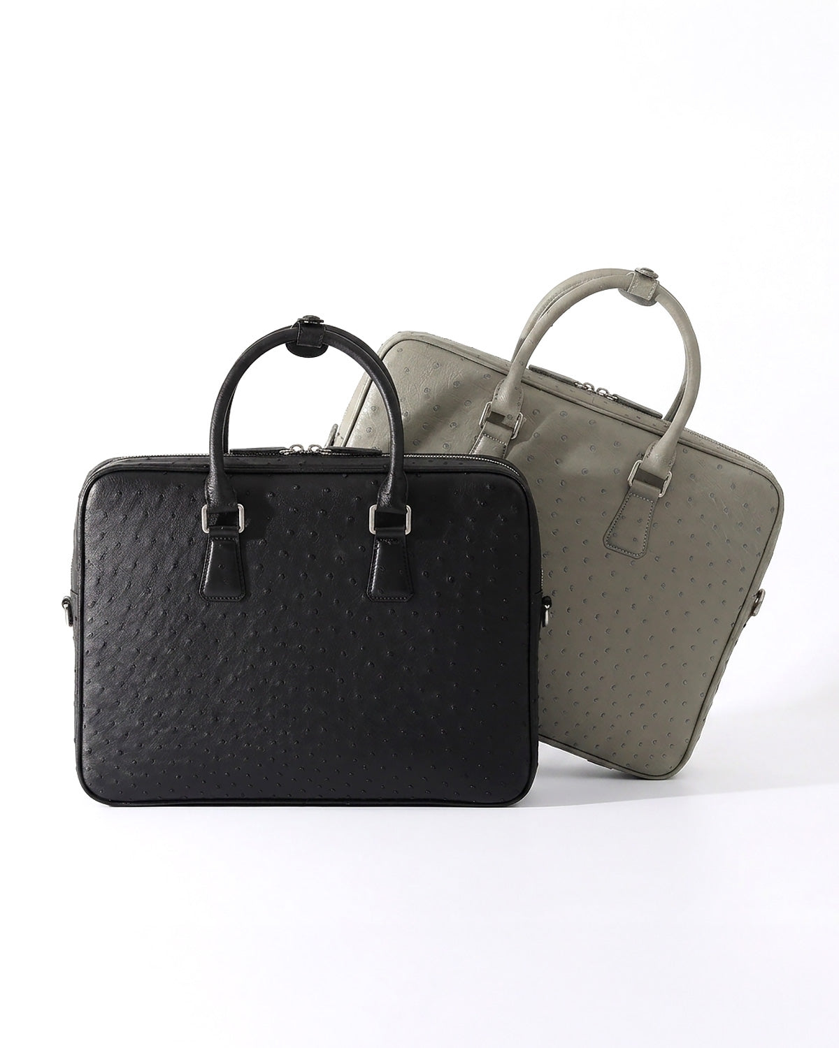 Two structured leather briefcases with top handles, shown on a clean white studio background. The front bag is a black genuine ostrich briefcase, and the rear bag is an elephant gray genuine ostrich briefcase | BUCKLE Collective