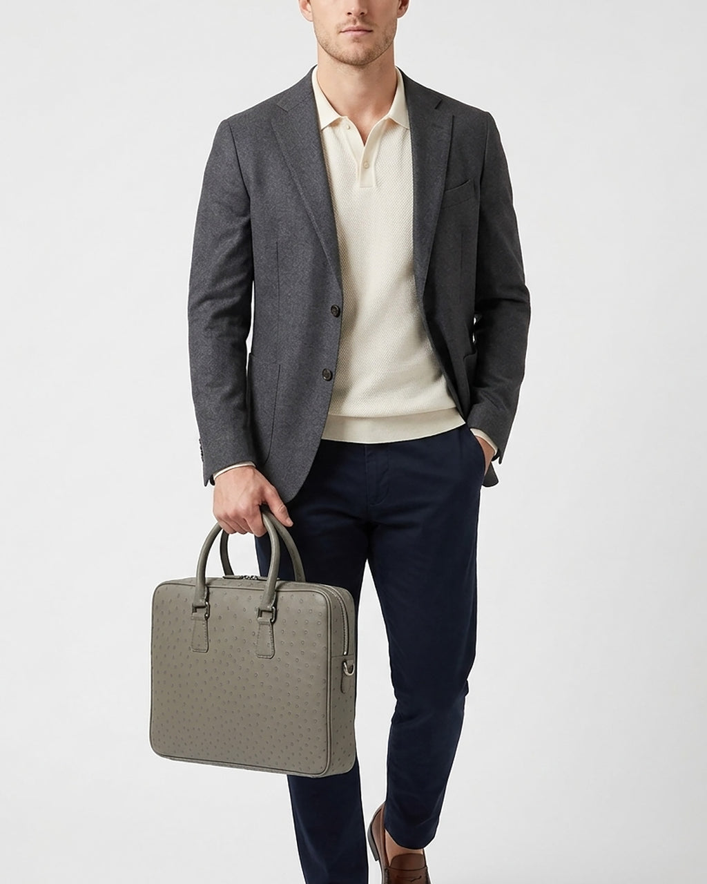 A man wearing a dark gray blazer, cream polo shirt, navy trousers, and brown shoes holds an elephant gray genuine ostrich leather briefcase | BUCKLE Collective