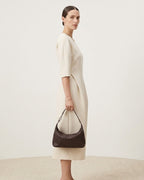 A woman stands in profile wearing a cream midi dress and holding a woven leather shoulder bag in coffee color