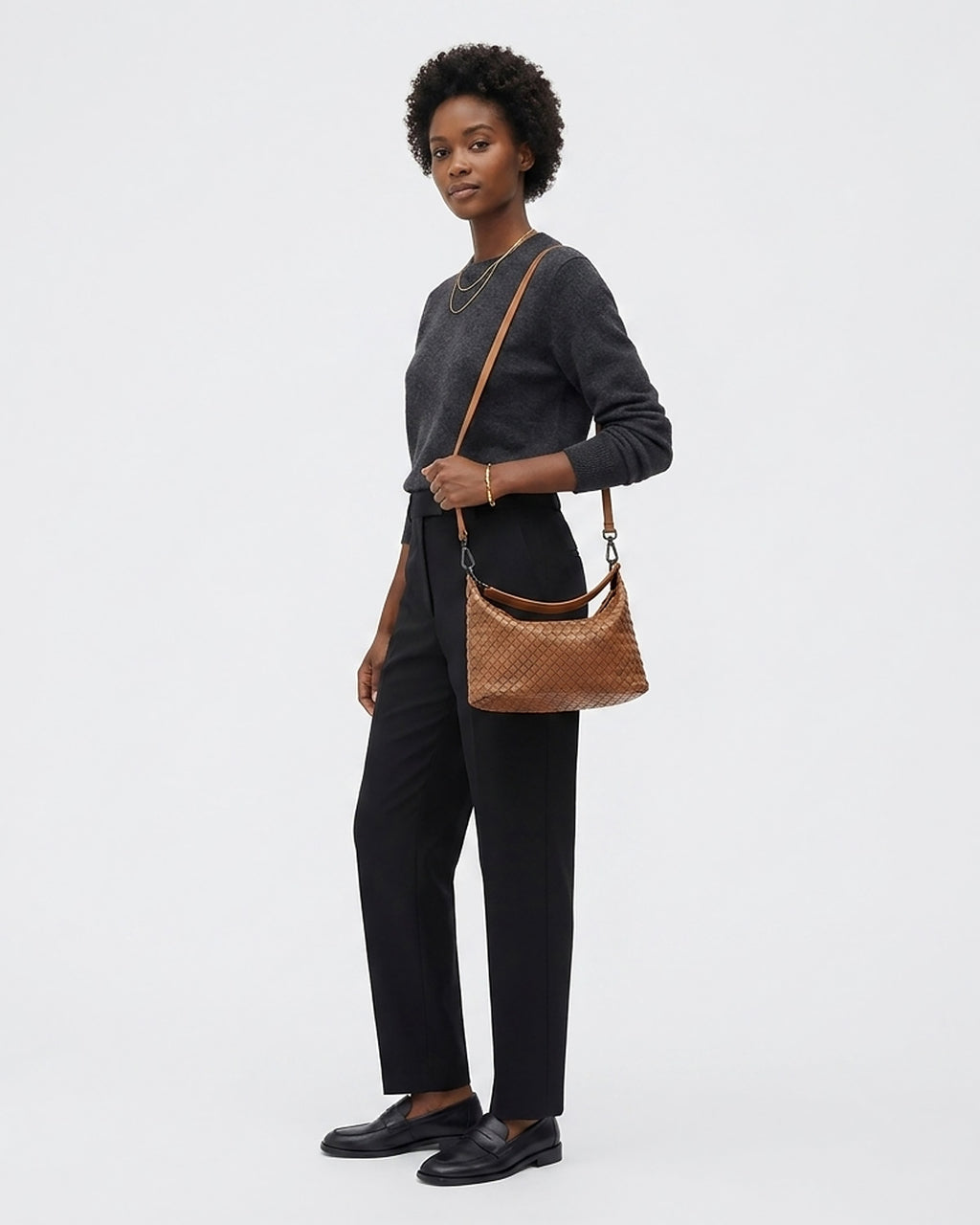 A woman stands against a light gray studio background wearing a dark charcoal long-sleeve top, black wide-leg trousers, black loafers, and a brown woven crossbody bag with a detachable strap.