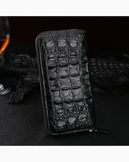 A black zip-around Genuine Siamese Crocodile Leather Clutch, displayed on a dark textured surface with a glass of whiskey and a black crocodile skin runner in the background.