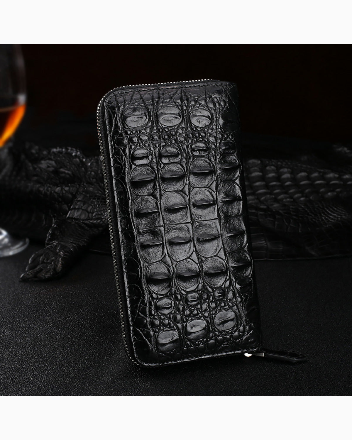 A black zip-around Genuine Siamese Crocodile Leather Clutch, displayed on a dark textured surface with a glass of whiskey and a black crocodile skin runner in the background.