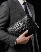 A man in a gray suit and tie holding a Black Genuine Siamese Crocodile Leather Clutch with a top zipper and a matching wrist strap