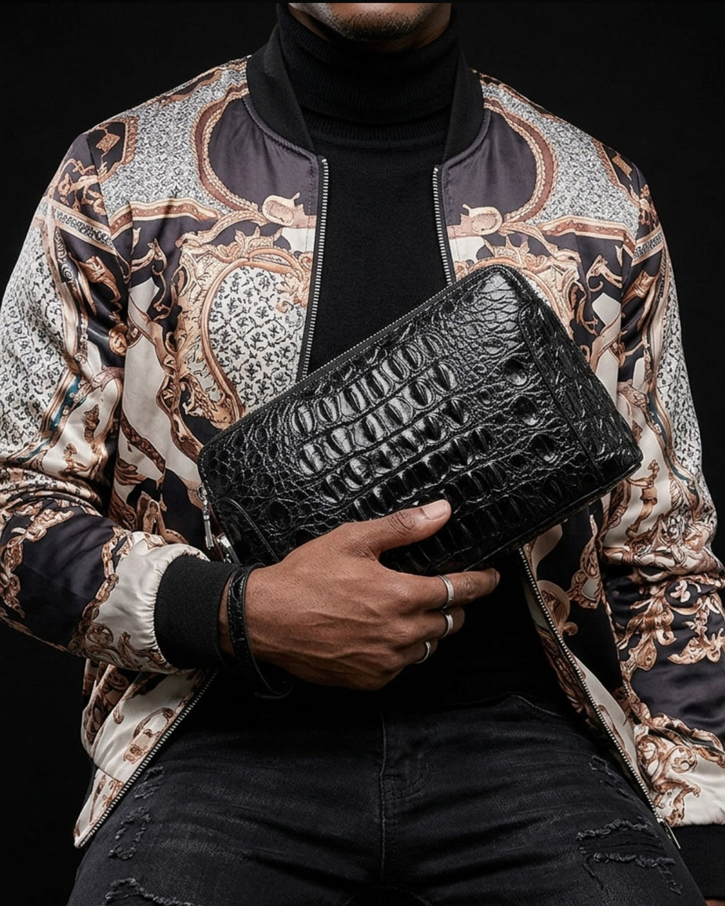 A man wearing a black turtleneck and a patterned satin bomber jacket holds a black crocodile leather clutch bag