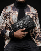 A man wearing a black turtleneck and a patterned satin bomber jacket holds a black crocodile leather clutch bag