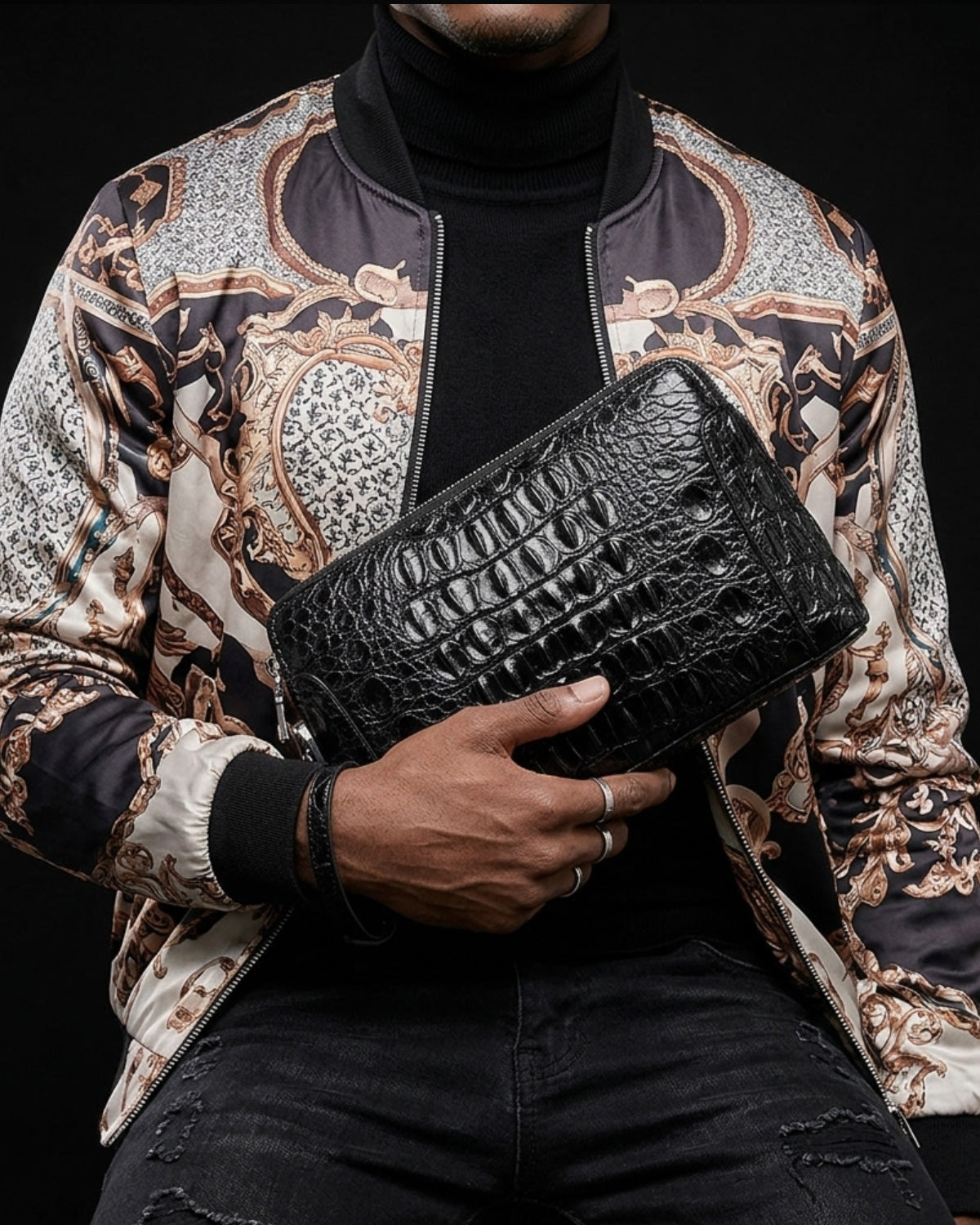 A man wearing a black turtleneck and a patterned satin bomber jacket holds a black crocodile leather clutch bag