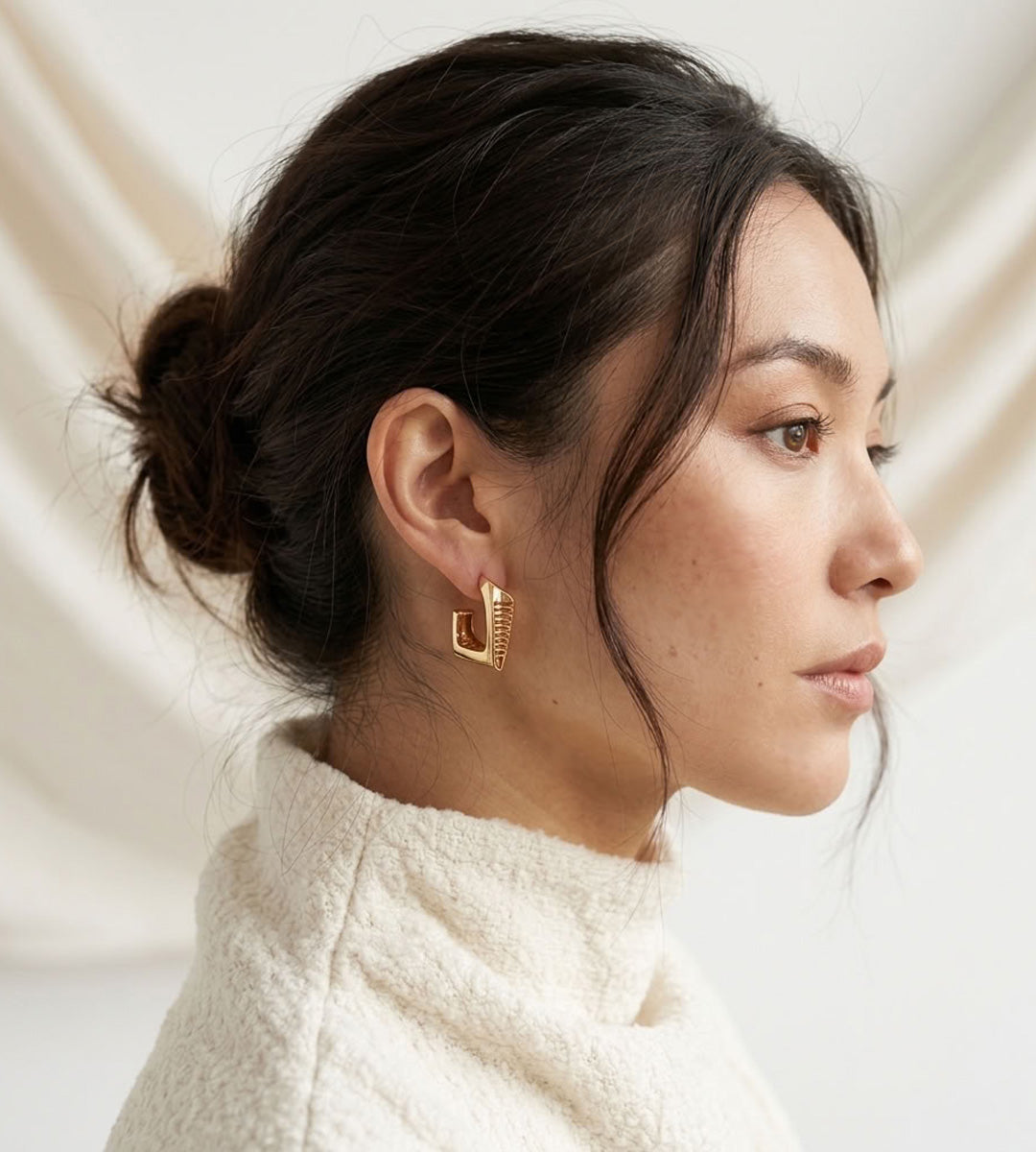 Side view of a model wearing a gold-tone angular hoop earring with ribbed detailing, styled with an ivory high-neck top.