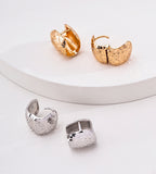 Four textured 18K gold-plated S925 Silver hoop-style earrings displayed on a white surface, including two gold-toned earrings and two silver-toned earrings, with one pair resting on a white curved platform and the other pair below it. BUCKLE Collective