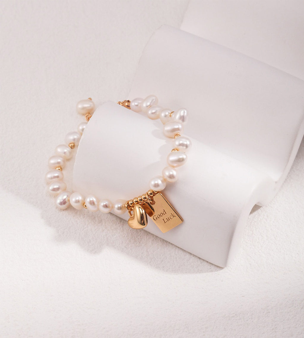 Good Luck baroque pearl bracelet worn on wrist in vintage gold — "Good Luck" tag and heart charm detail, styled with gold rings and pearl necklace, 18K gold plated S925 silver, BUCKLE Collective

